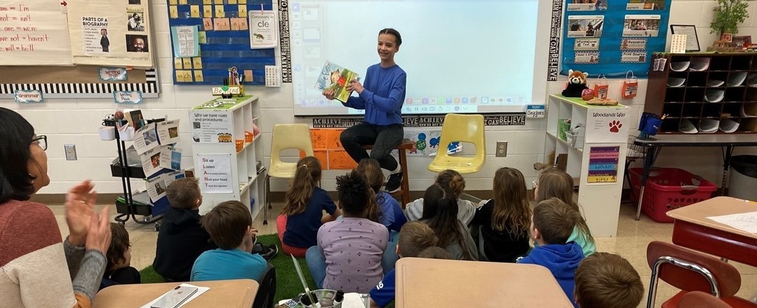 Students sitting listening to an older student read a book
